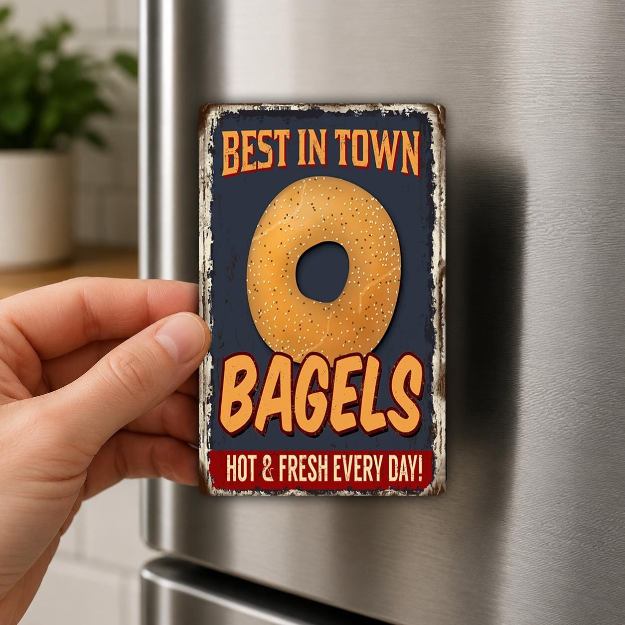A hand is holding a metal magnet with a picture of a bagel and text that says "Best in Town BAGELS HOT & FRESH EVERY DAY!"