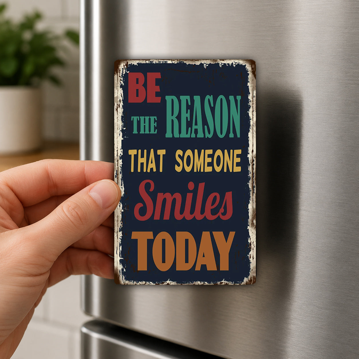 A hand holding a metal sign with the text "BE THE REASON THAT SOMEONE SMILES TODAY" on it.