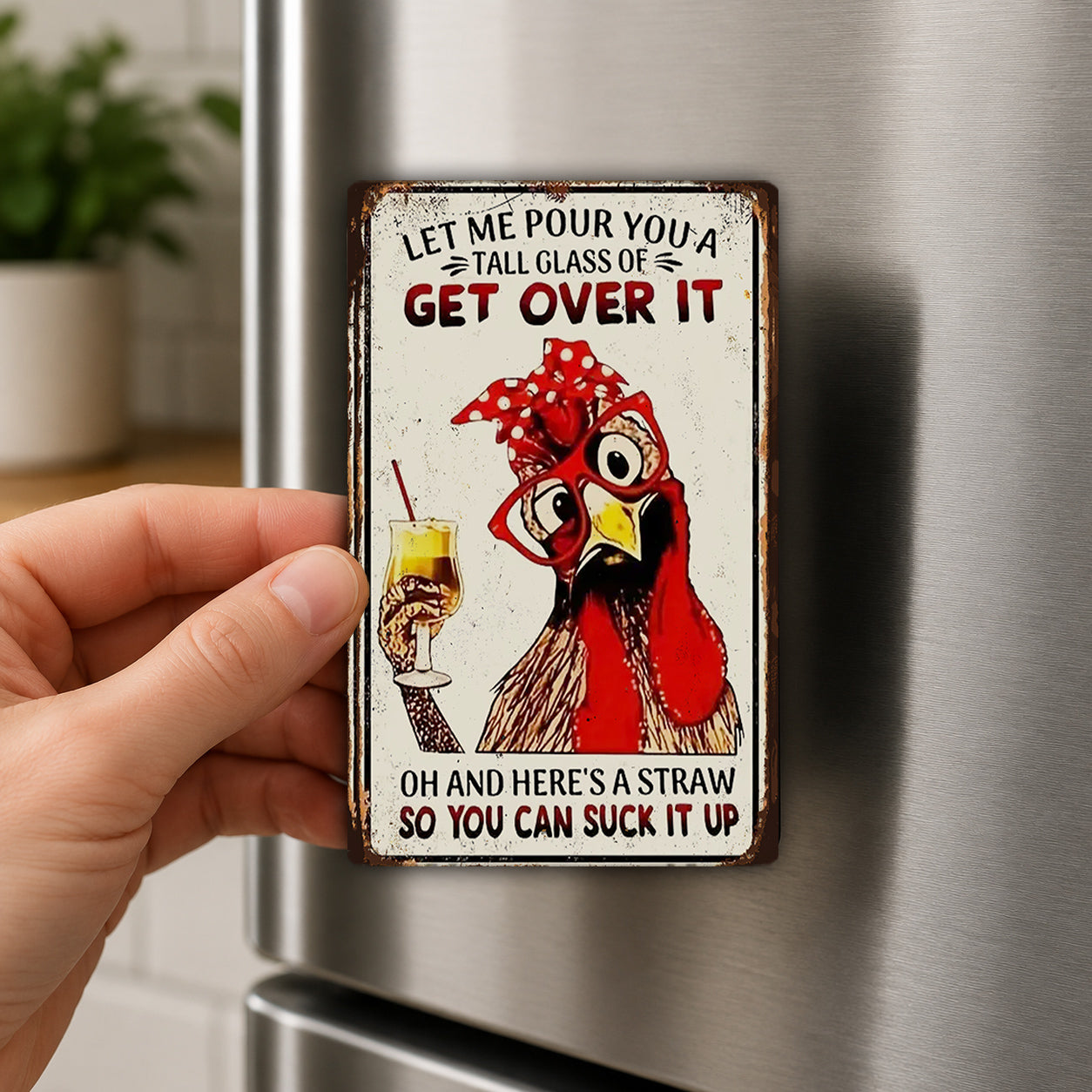 A person's hand is holding a metal sign with a cartoon image of a chicken and text that says "Let me pour you a tall glass of get over it".
