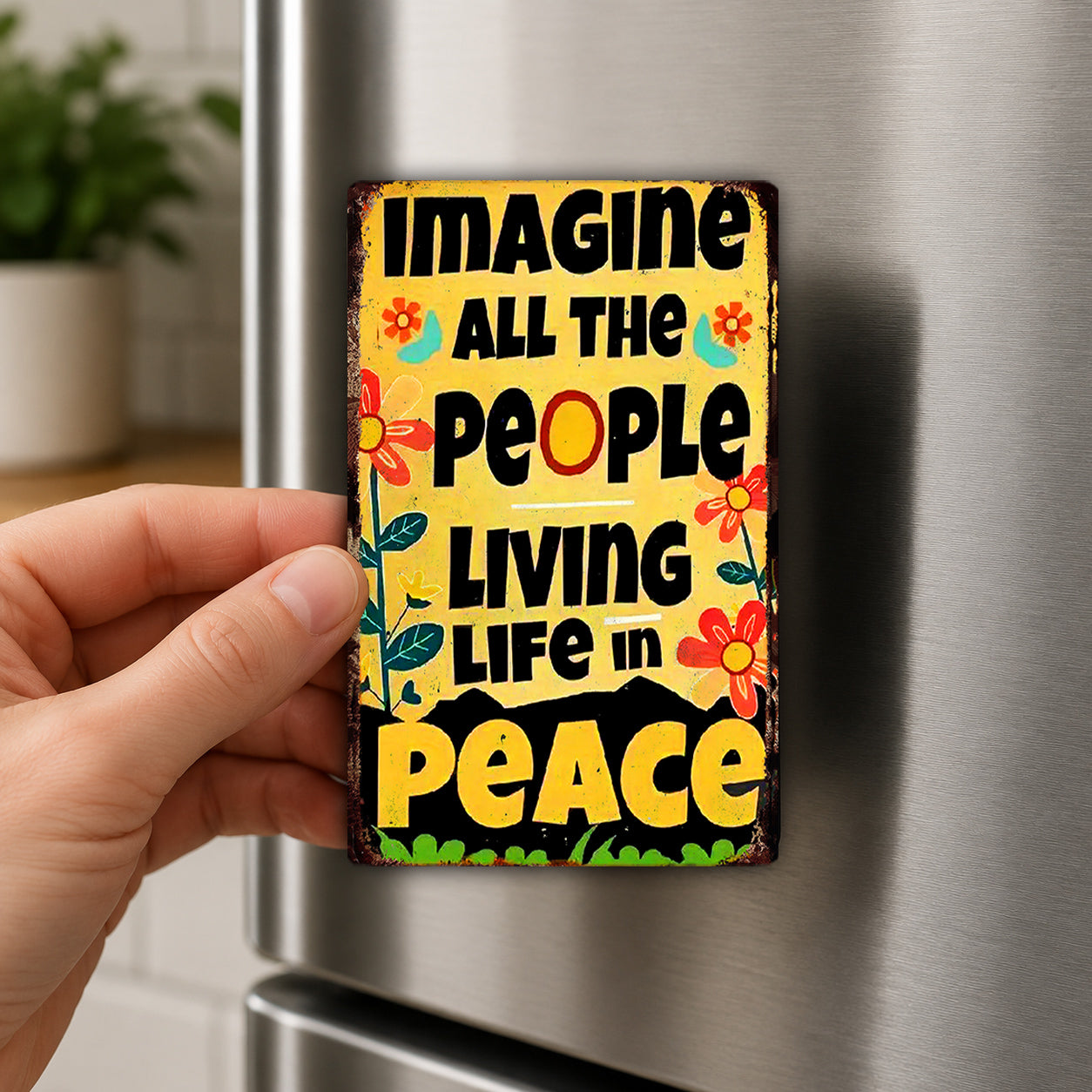 A hand is holding a colorful, vintage-style magnet with the text "IMAGINE ALL THE PEOPLE LIVING LIFE IN PEACE" on it.