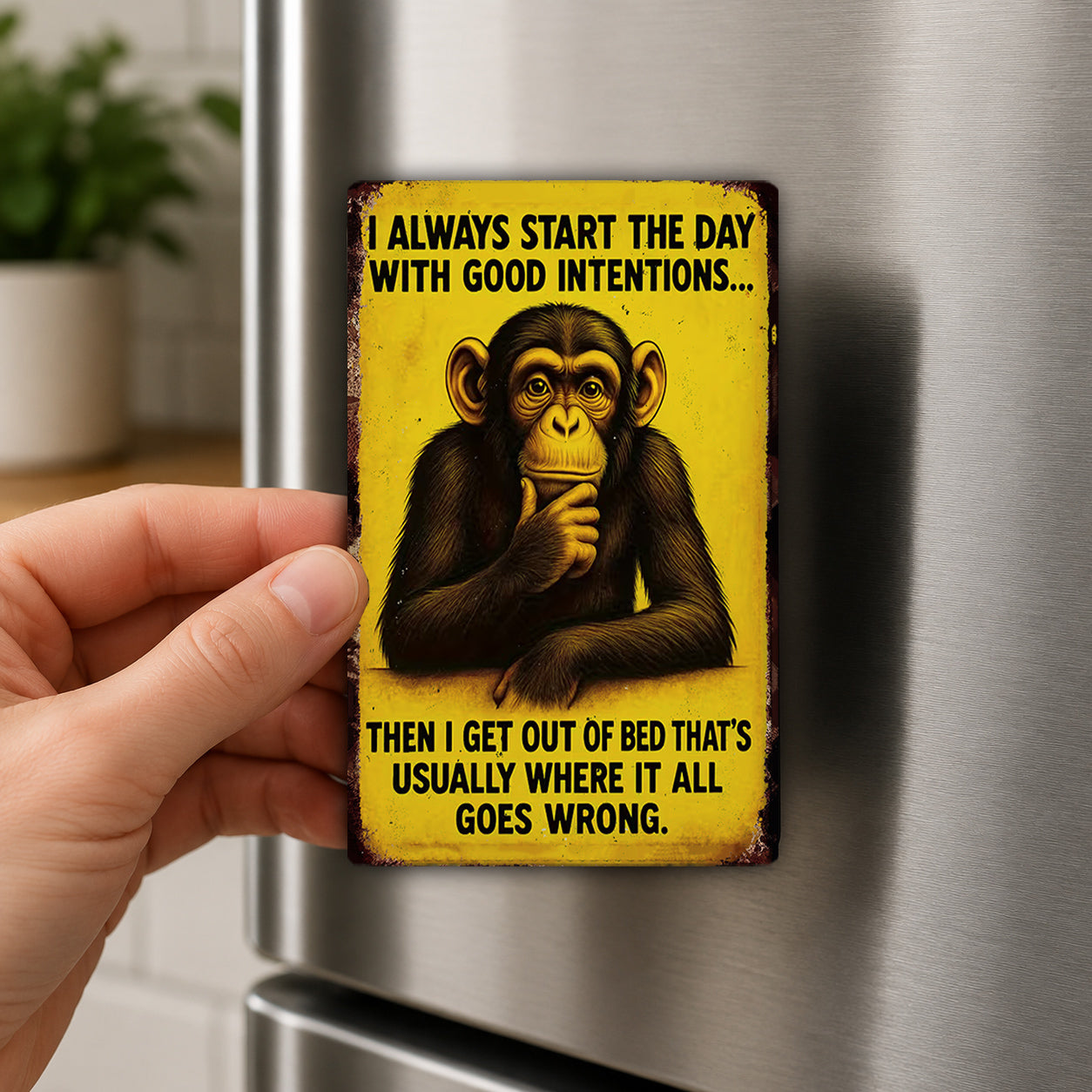 A person is holding a yellow magnet with a cartoon image of a monkey and a humorous text that reads "I always start the day with good intentions... then I get out of bed that's usually where it all goes wrong."