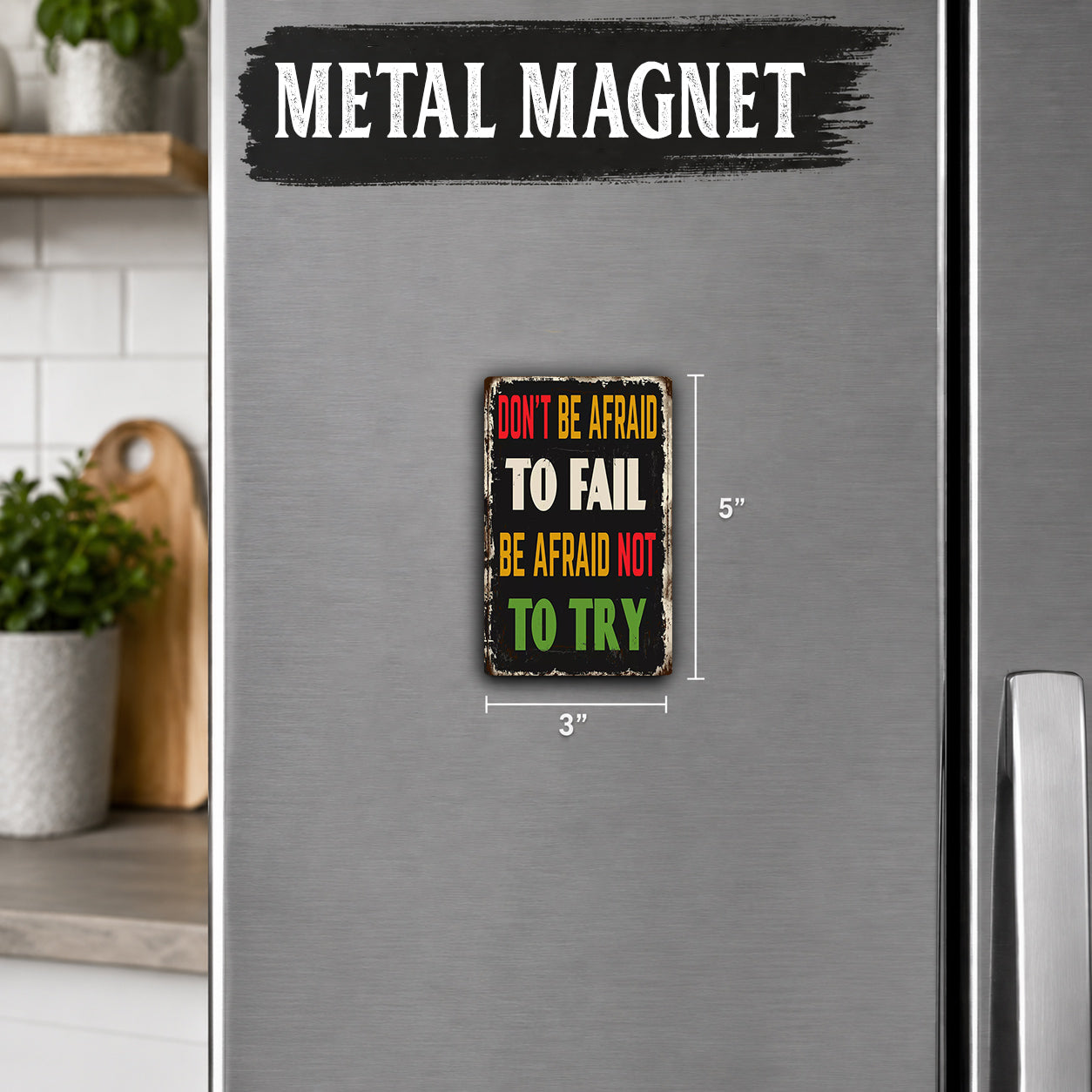 a small, vintage-style metal magnet with the text "Don't be afraid to fail. Be afraid not to try." The magnet is placed on a refrigerator, which is part of a kitchen setting.