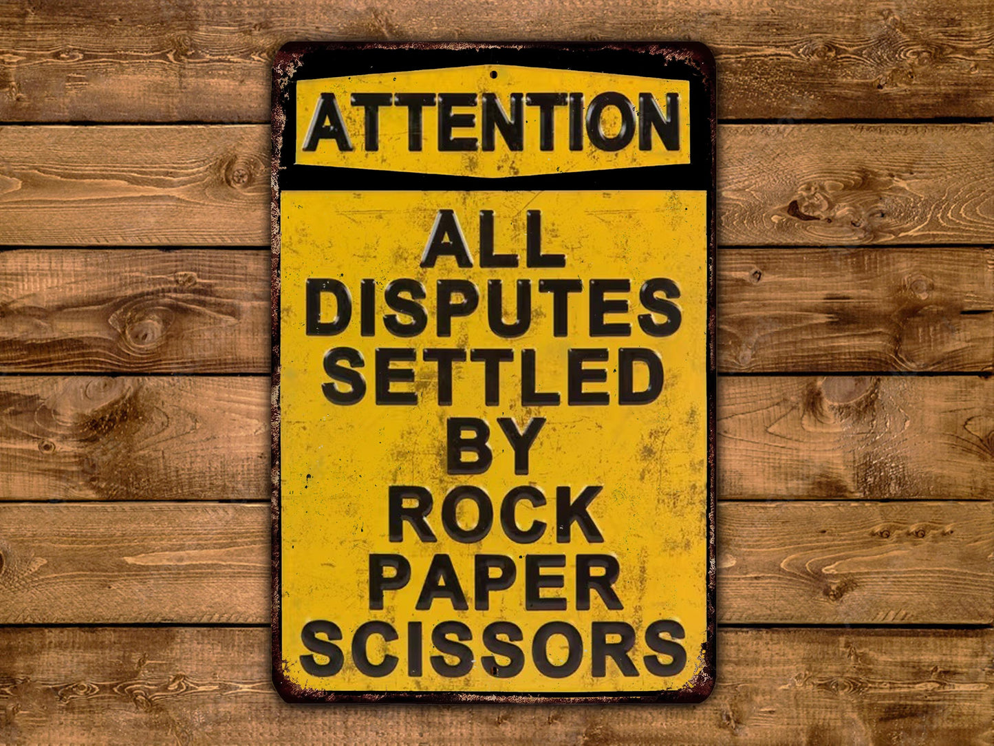 A yellow sign with black text that reads "ATTENTION ALL DISPUTES SETTLEED BY ROCK PAPER SCISSORS" is displayed on a wooden background.