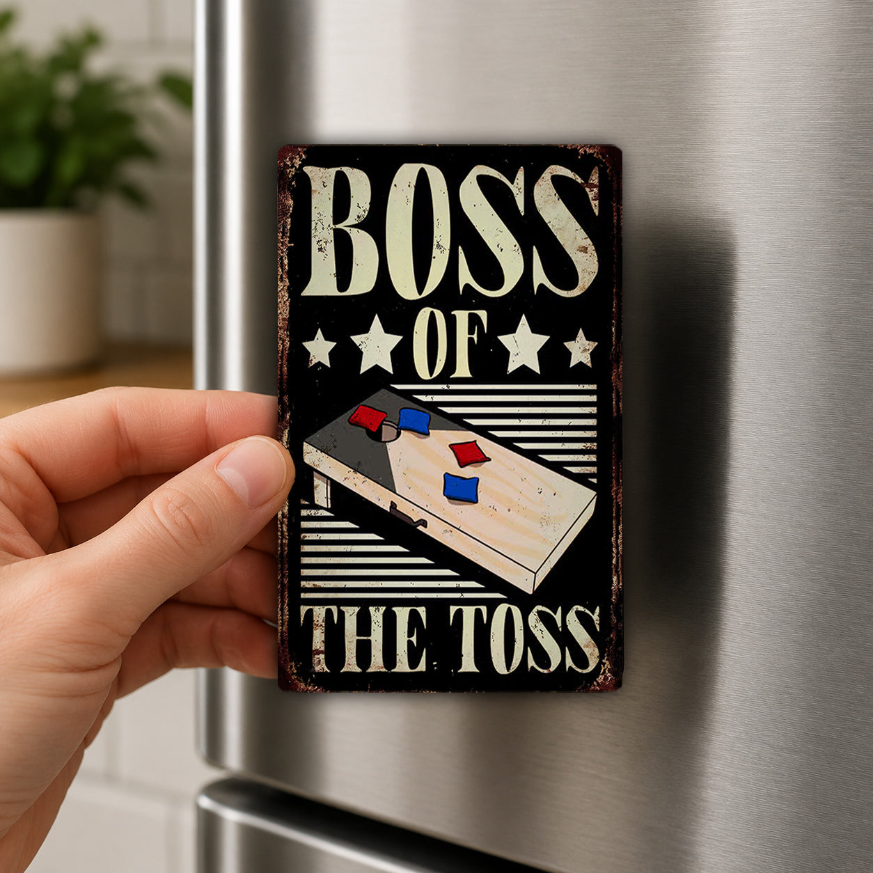 A person's hand is holding a metal object with the text "BOSS OF THE TOSS" and an illustration of a cornhole board.