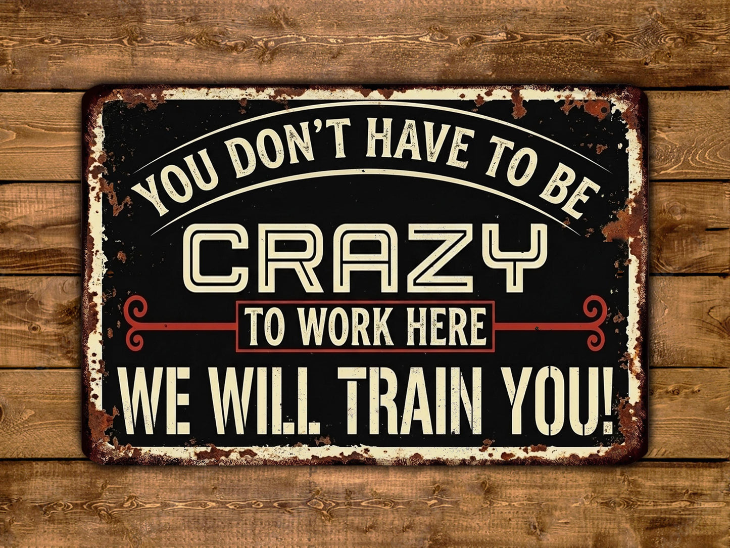 A vintage-style metal sign with the text "YOU DON'T HAVE TO BE CRAZY TO WORK HERE WE WILL TRAIN YOU!" displayed on a wooden background.
