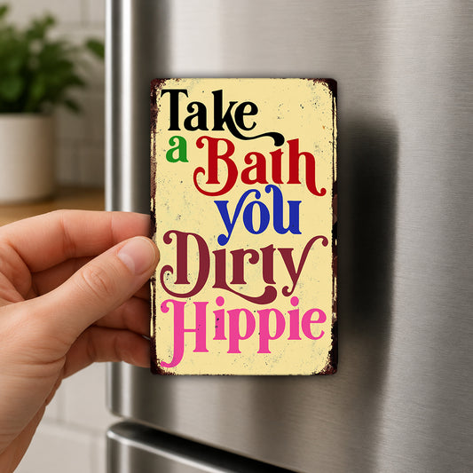 A hand is holding a colorful, vintage-style magnet with the text "Take a Bath You Dirty Hippie" on it.