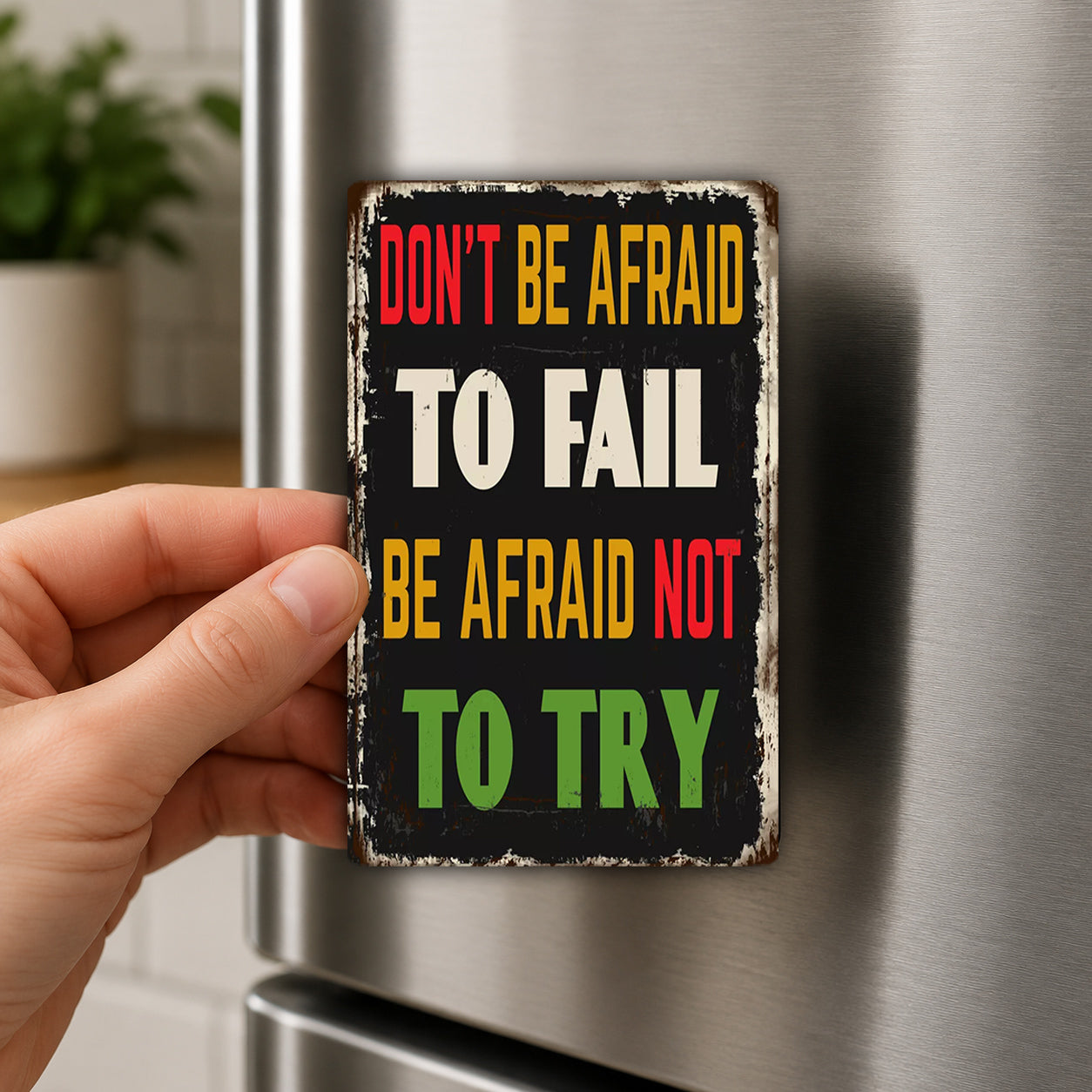 A person's hand is holding a metal sign with the text "DON'T BE AFRAID TO FAIL BE AFRAID NOT TO TRY" on it.