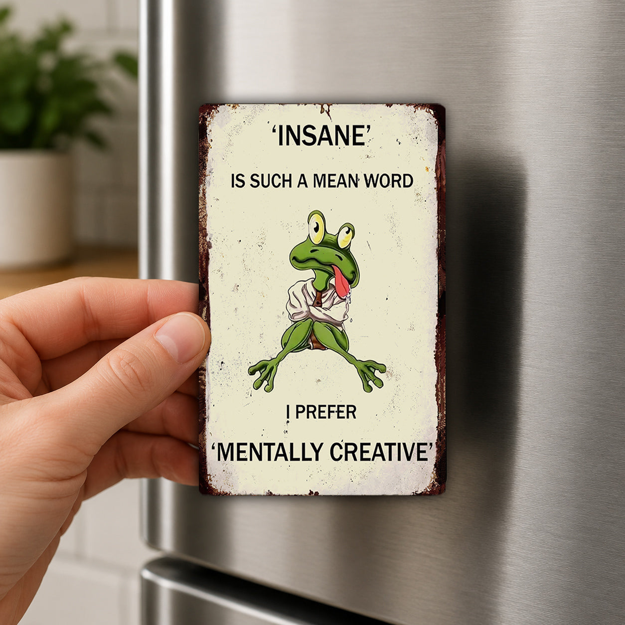 a person holding a small, vintage-style metal magnet with a cartoon frog on it. The magnet has text that reads "INSANE" and "I PREFER 'MENTALLY CREATIVE'".