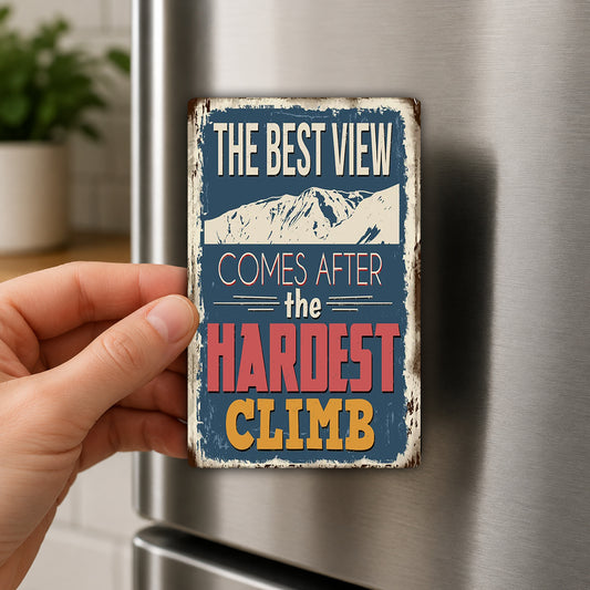 A hand is holding a metal refrigerator magnet with the text "THE BEST VIEW COMES AFTER THE HARDEST CLIMB" on it.