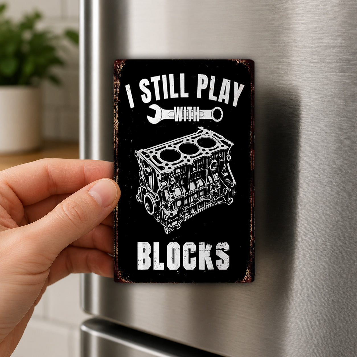 A person's hand is holding a black and white metal magnet with the text "I STILL PLAY WITH BLOCKS" and an illustration of a car engine.