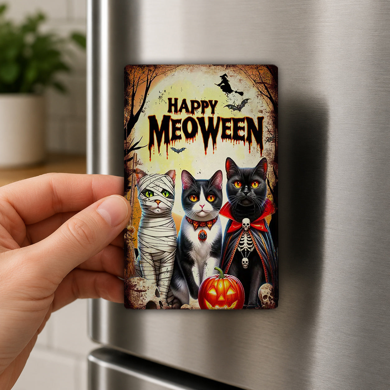A hand is holding a Halloween-themed magnet featuring three cats dressed in costumes, with a pumpkin and bats in the background.