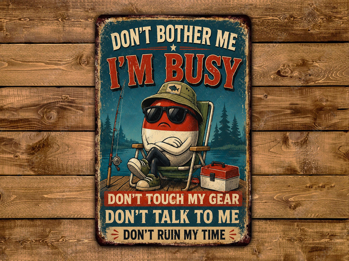 a vintage-style sign with a cartoon character sitting in a chair, fishing, and wearing sunglasses. The sign has a humorous message that reads "Don't bother me, I'm busy, don't touch my gear, don't talk to me, don't ruin my time".