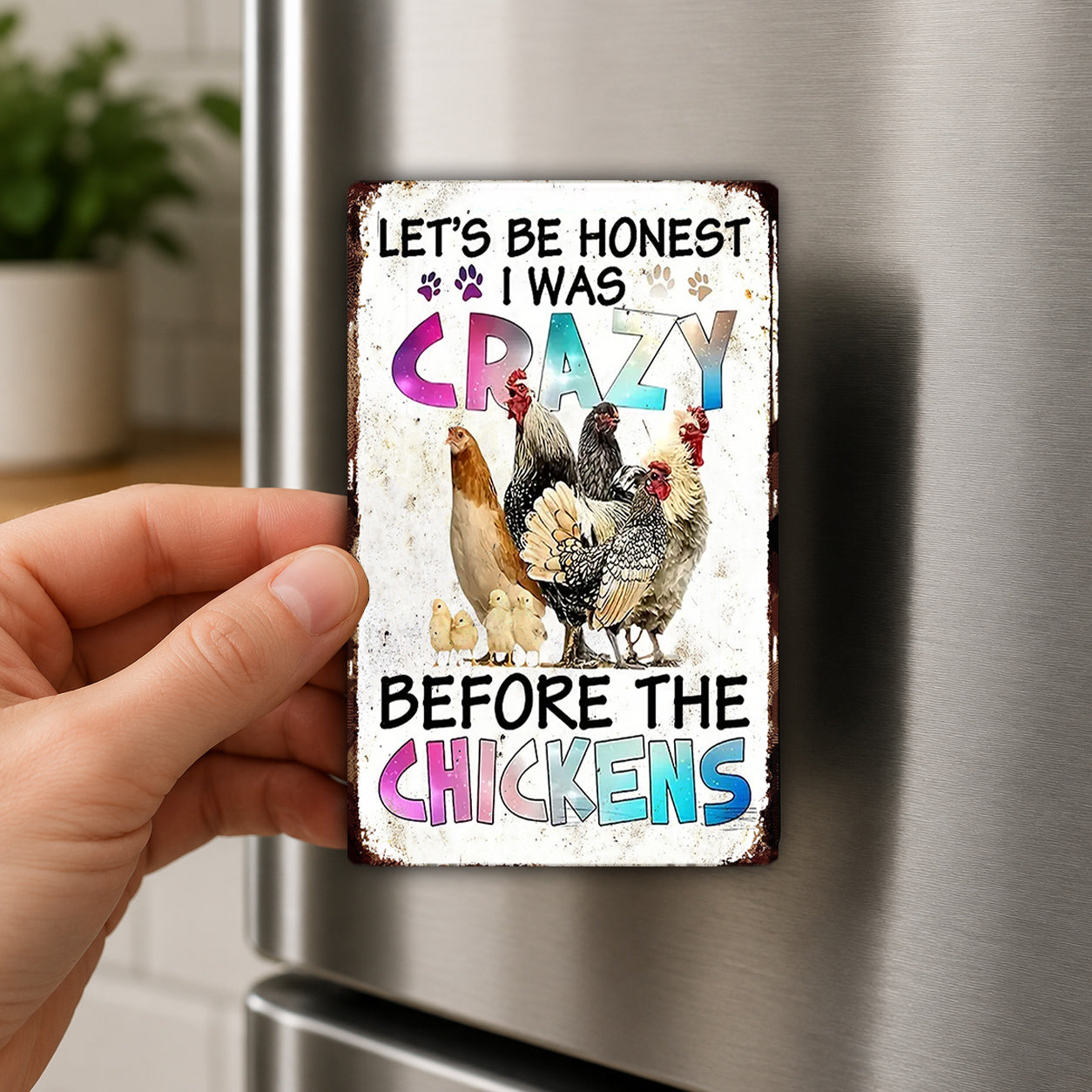 A person's hand holding a metal refrigerator magnet with a colorful design featuring chickens and text.