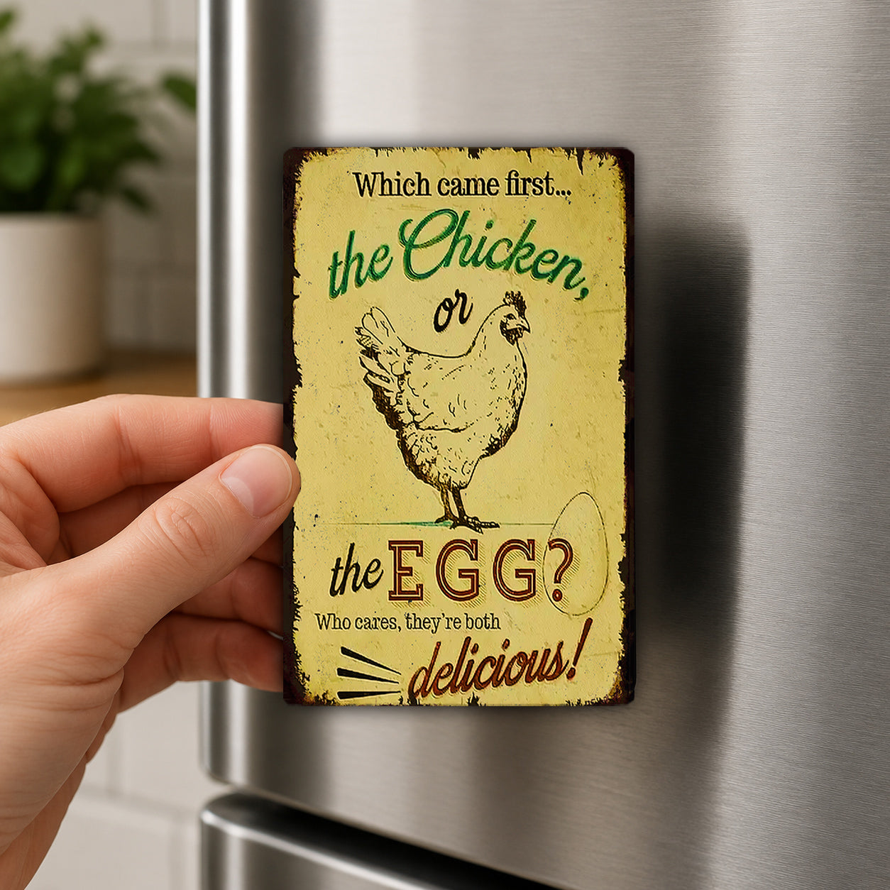 A person's hand is holding a vintage-style magnet with a humorous message about chickens and eggs.