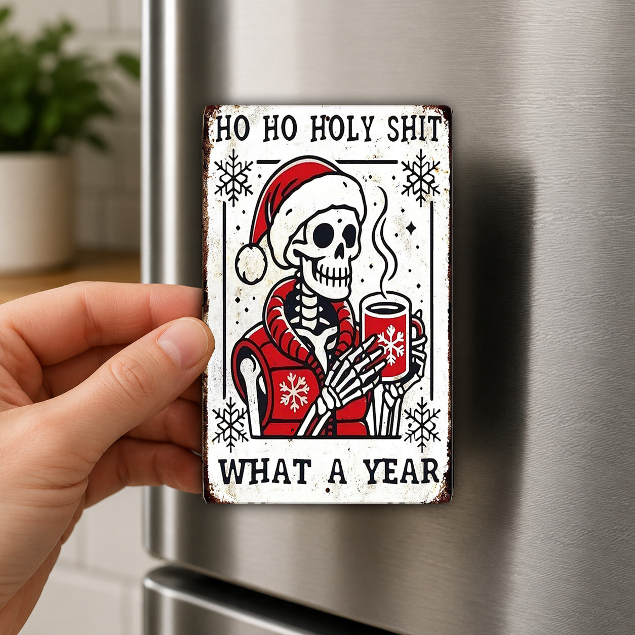 A person's hand is holding a metal magnet with a cartoon skeleton wearing a Santa hat and holding a mug, with the text "HO HO HOLY SHIT WHAT A YEAR" written above and below the image.