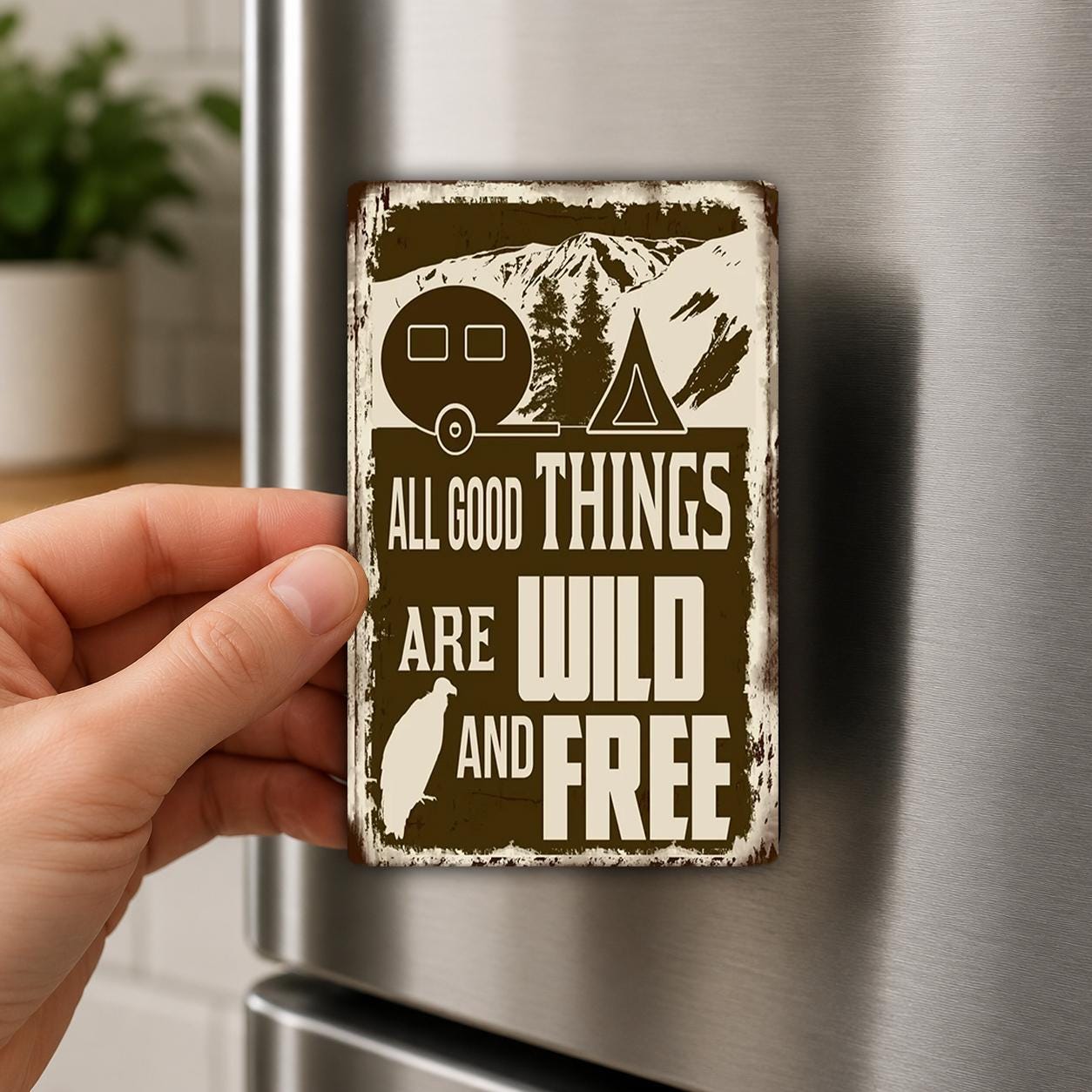 A hand is holding a metal magnet with the text "ALL GOOD THINGS ARE WILD AND FREE" on it.