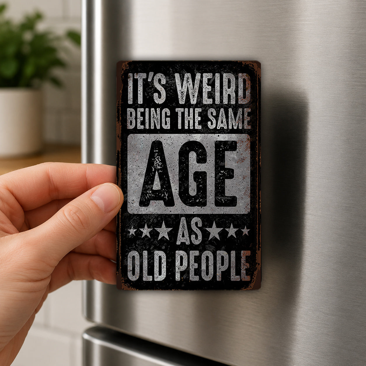 A person's hand is holding a metal sign with the text "IT'S WEIRD BEING THE SAME AGE AS OLD PEOPLE" on it.