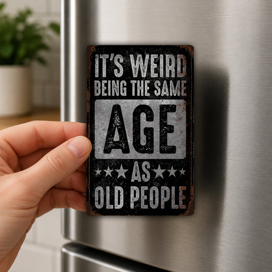 A person's hand is holding a metal sign with the text "IT'S WEIRD BEING THE SAME AGE AS OLD PEOPLE" on it.