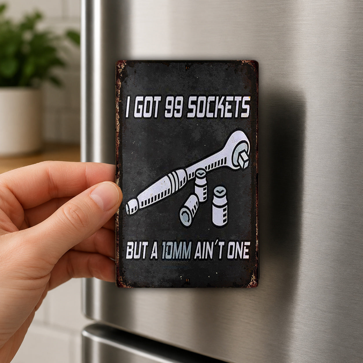 A person's hand is holding a metal magnet with a humorous text and an illustration on it, which appears to be a humorous take on the phrase "99 sockets but a 10mm ain't one".