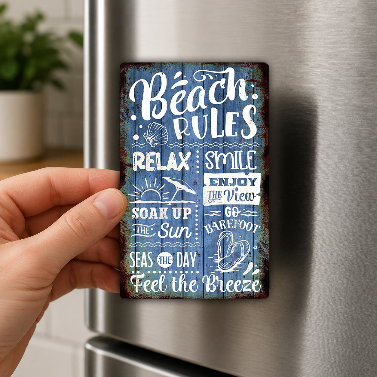 A hand is holding a metal sign with text on it, which appears to be a beach-themed decoration or magnet.