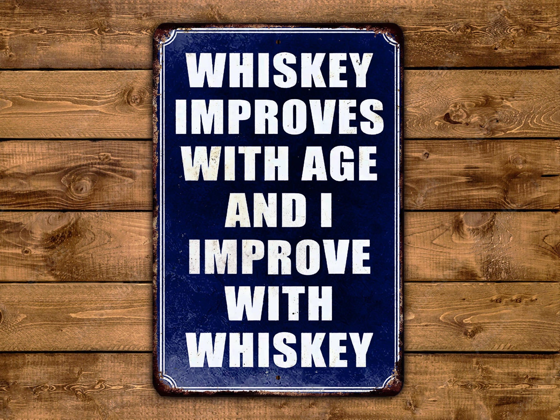 A blue and white sign with the text "WHISKEY IMPROVES WITH AGE AND I IMPROVE WITH WHISKEY" displayed on a wooden background.