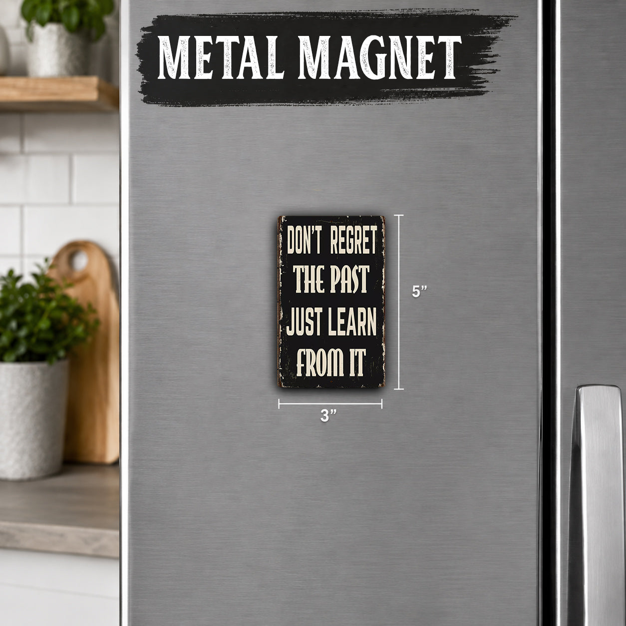 a small black and white metal magnet with the text "Don't Regret the Past Just Learn from It" on it. The magnet is placed on a refrigerator, and there is a wooden cutting board visible in the background.