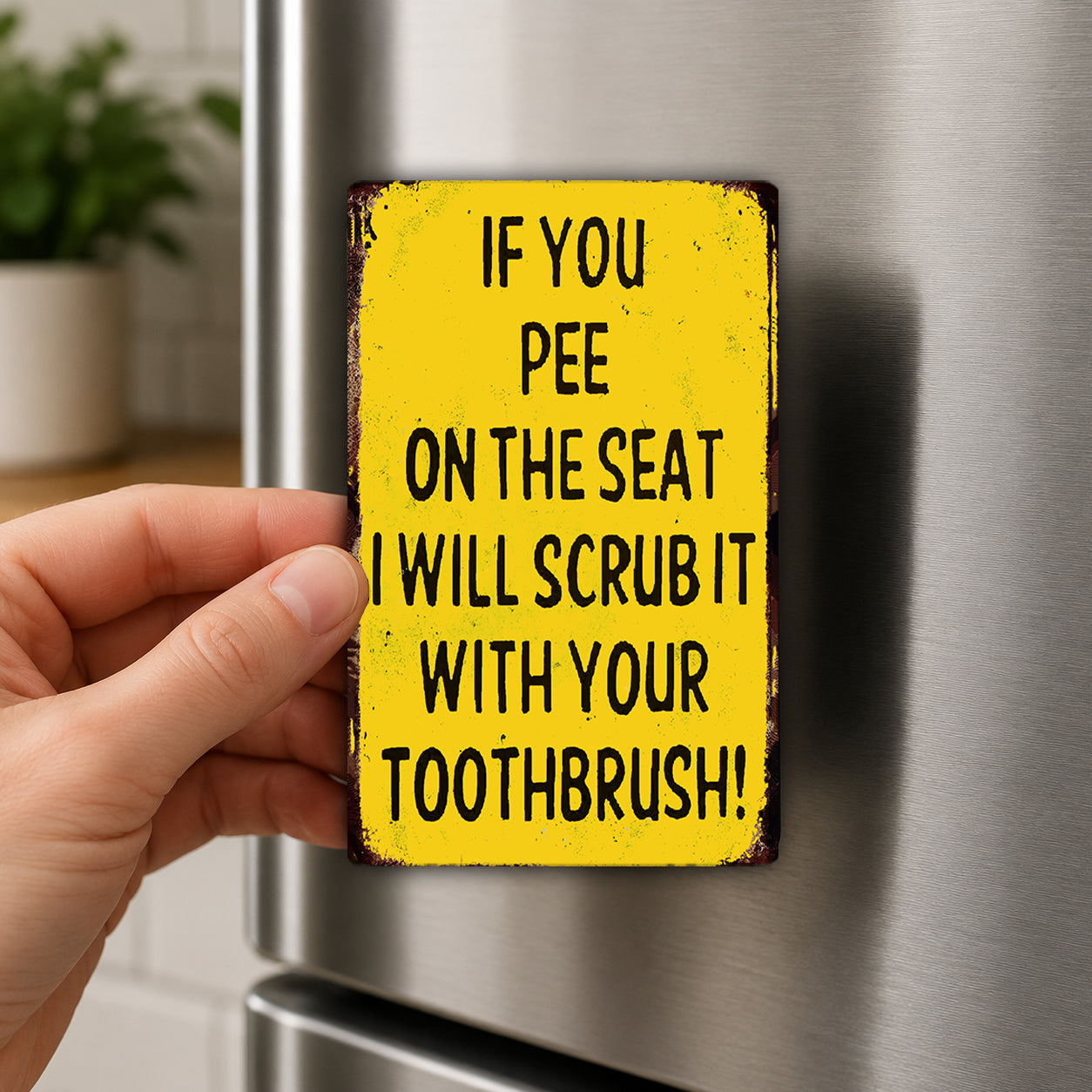 A person's hand holding a yellow sign with the text "IF YOU PEE ON THE SEAT I WILL SCRUB IT WITH YOUR TOOTHBRUSH!"
