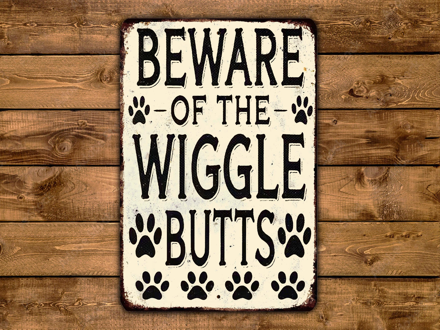 A metal sign with the text "BEWARE OF THE WIGGLES BUTTS" and paw prints, placed on a wooden background.