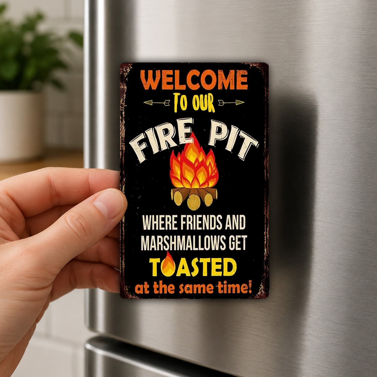 A person's hand holding a sign that says "Welcome to our fire pit" with a fire pit illustration and the text "Where friends and marshmallows get toasted at the same time!"