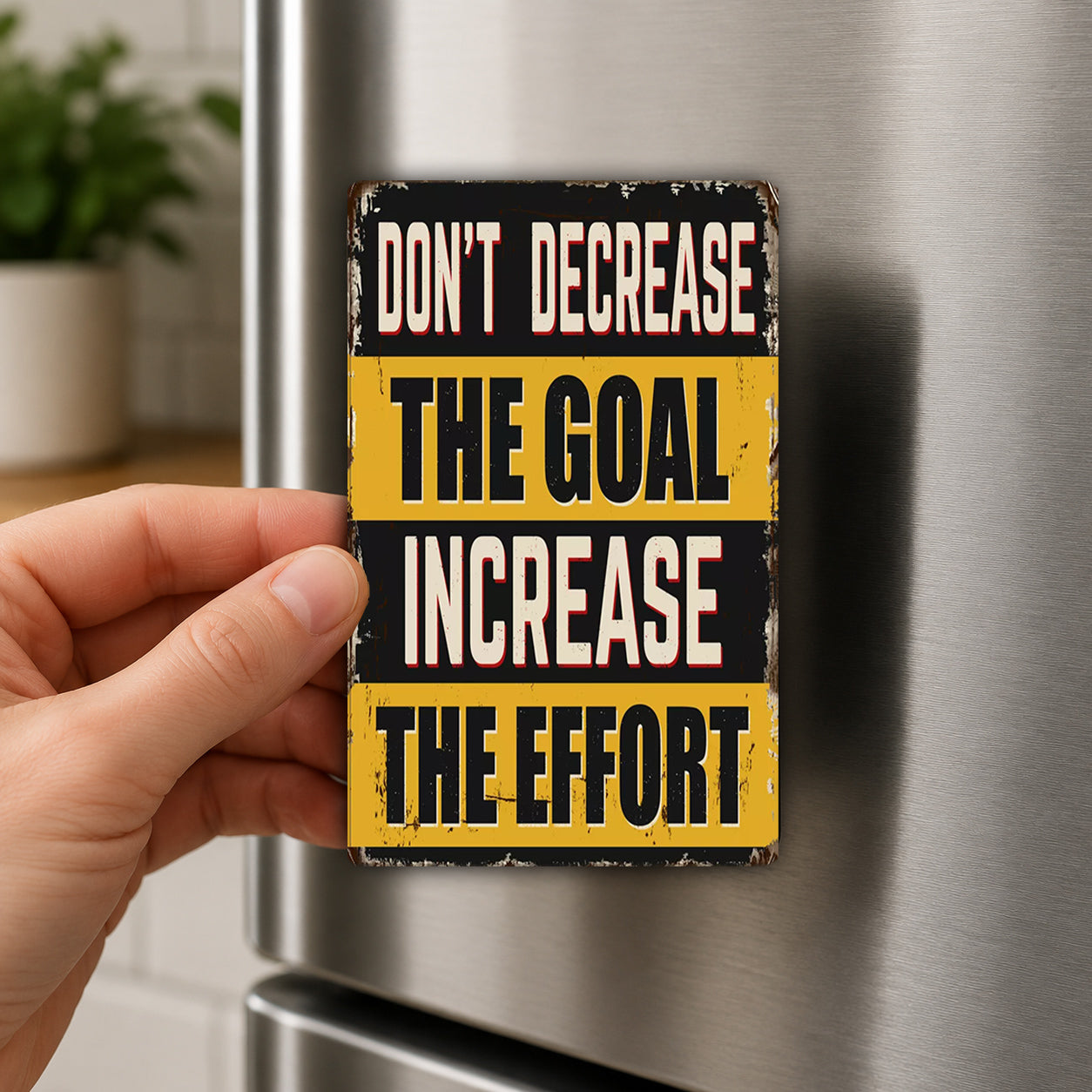 a person's hand holding a small, vintage-style metal magnet with the text "DON'T DECREASE THE GOAL INCREASE THE EFFORT" on it.