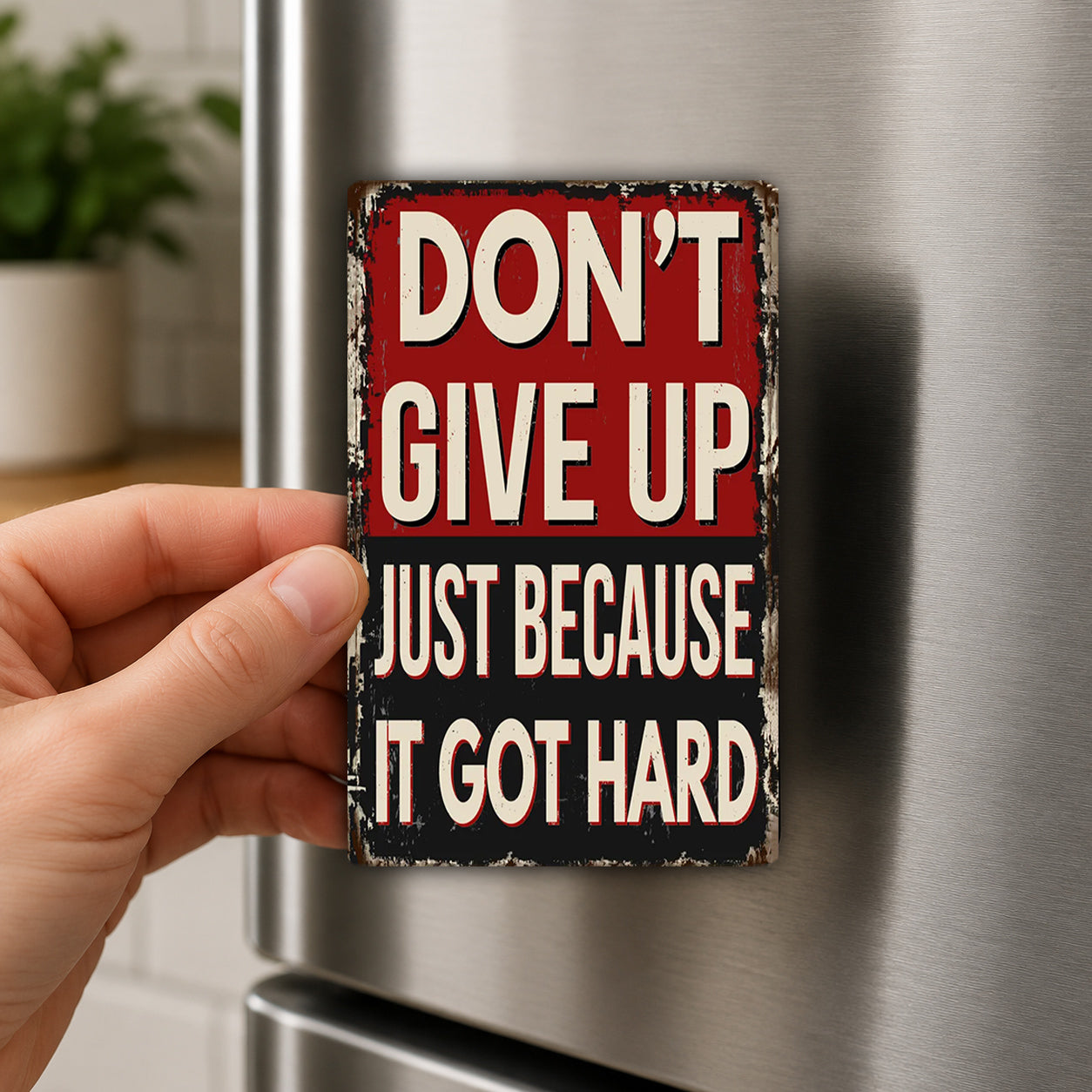 A person's hand is holding a small metal sign with the text "DON'T GIVE UP JUST BECAUSE IT GOT HARD" on it.