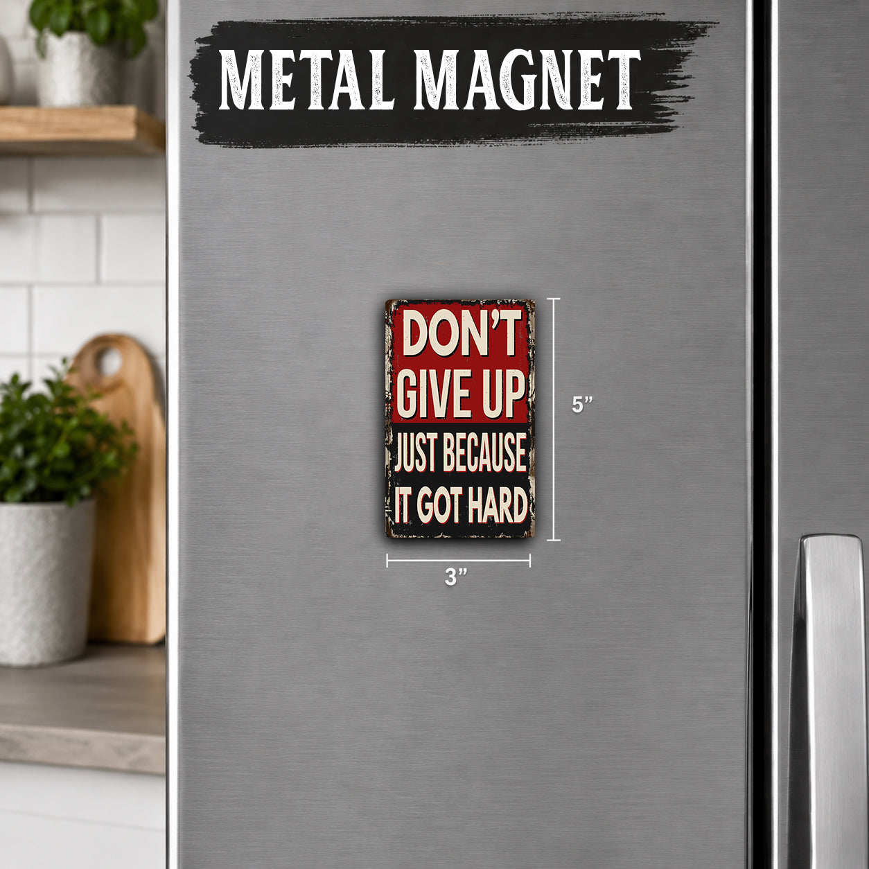 a small metal magnet with the text "DON'T GIVE UP" and "JUST BECAUSE IT GOT HARD" on it. The magnet is placed on a refrigerator, which is part of a kitchen setting.