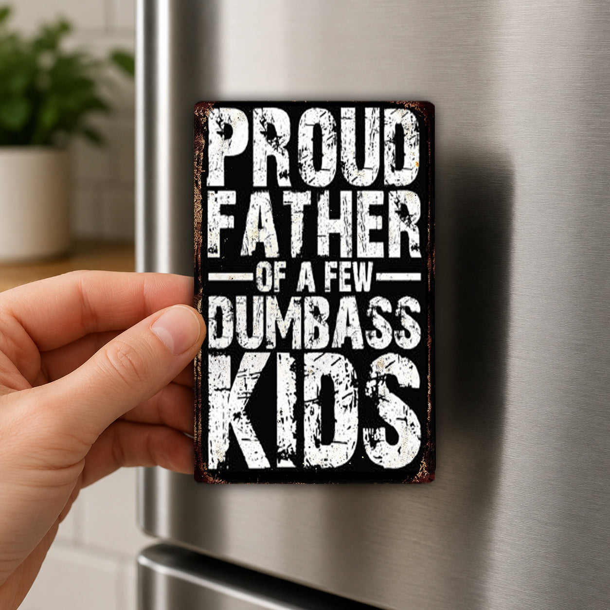 A hand holding a metal object with the text "Proud Father of a Few Dumbass Kids" written on it.