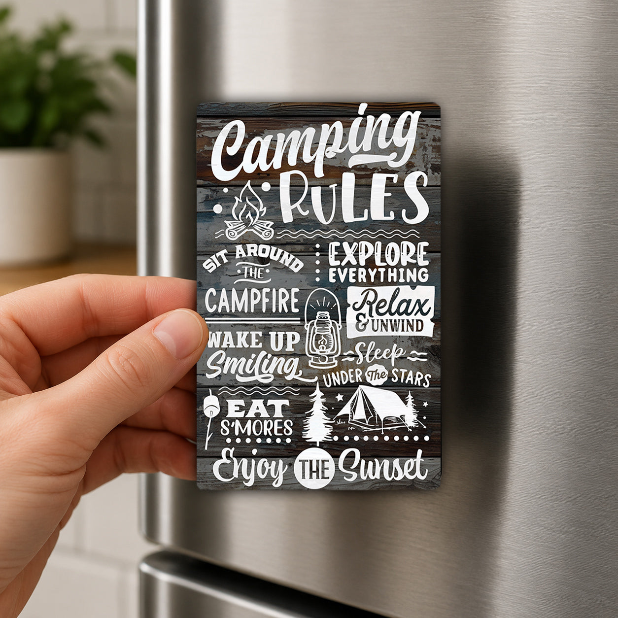 a hand holding a magnet with various camping-related phrases and illustrations on it.