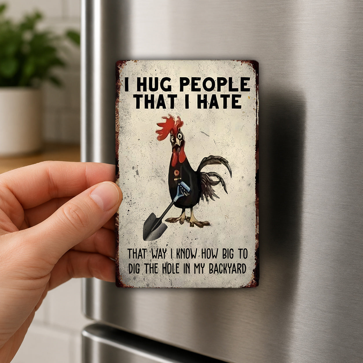 A person's hand is holding a metal refrigerator magnet with a cartoon rooster and the text "I HUG PEOPLE THAT I HATE" and "THAT WAY I KNOW HOW BIG TO DIG THE HOLE IN MY BACKYARD".