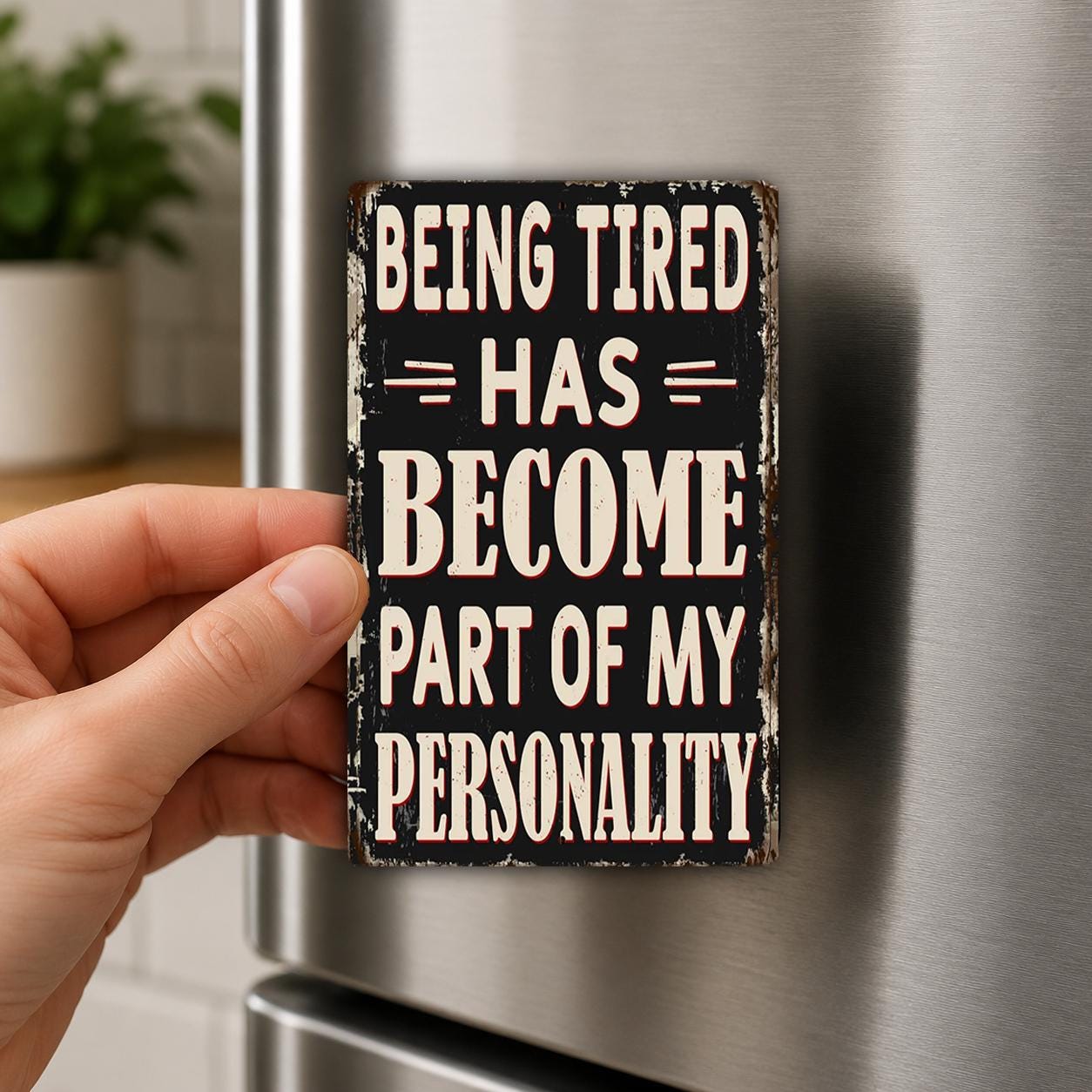 A person's hand is holding a sign with the text "Being Tired Has Become Part of My Personality" against a metallic background, which appears to be a refrigerator.