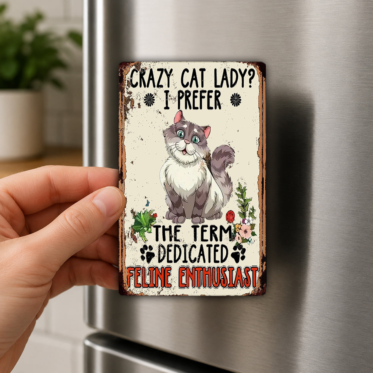 a person holding a small, vintage-style magnet with a cartoon cat on it. The magnet has text that reads "Crazy Cat Lady? I Prefer The Term Dedicated Feline Enthusiast" and features a cat illustration.