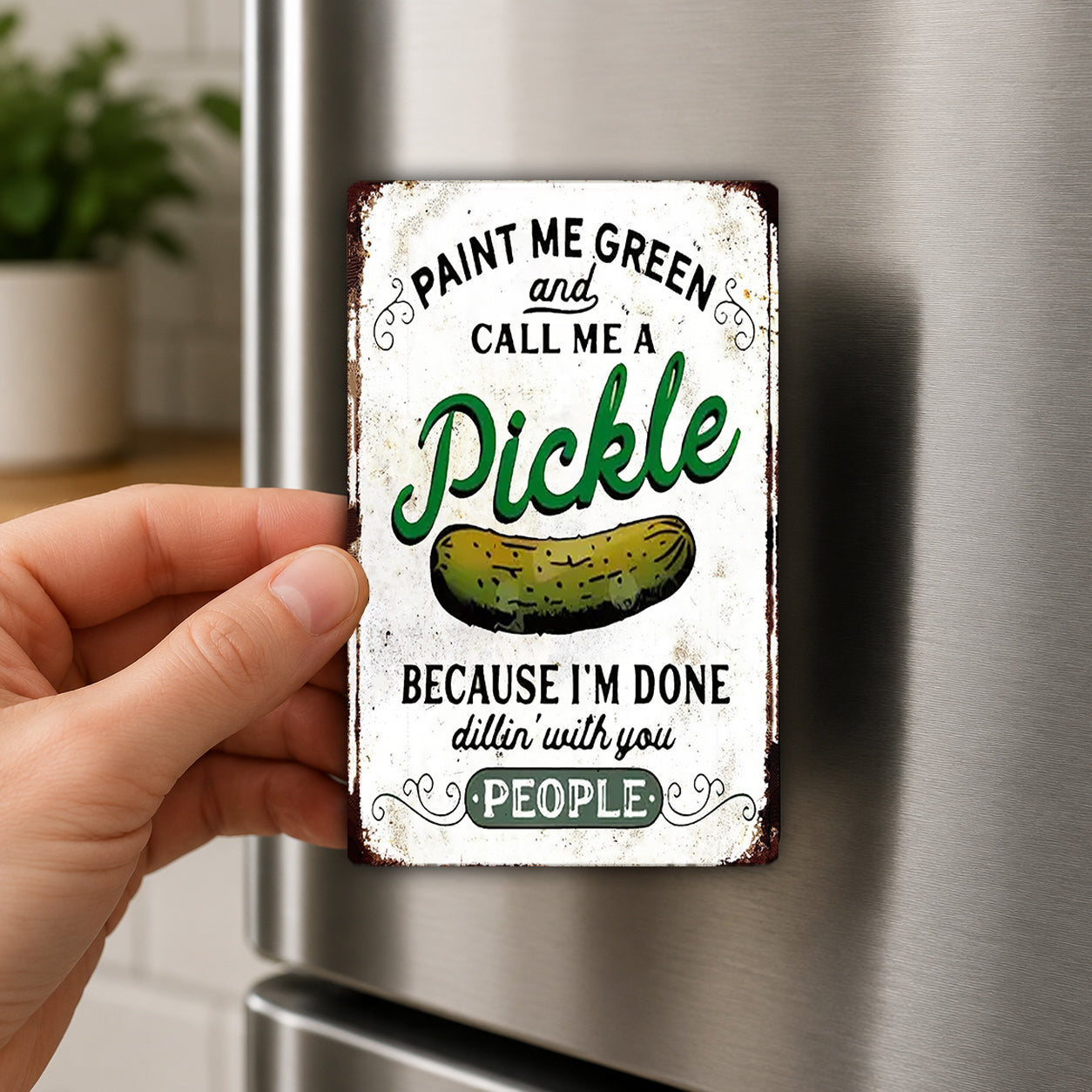 A person's hand is holding a metal magnet with a humorous text and an illustration of a pickle on it.