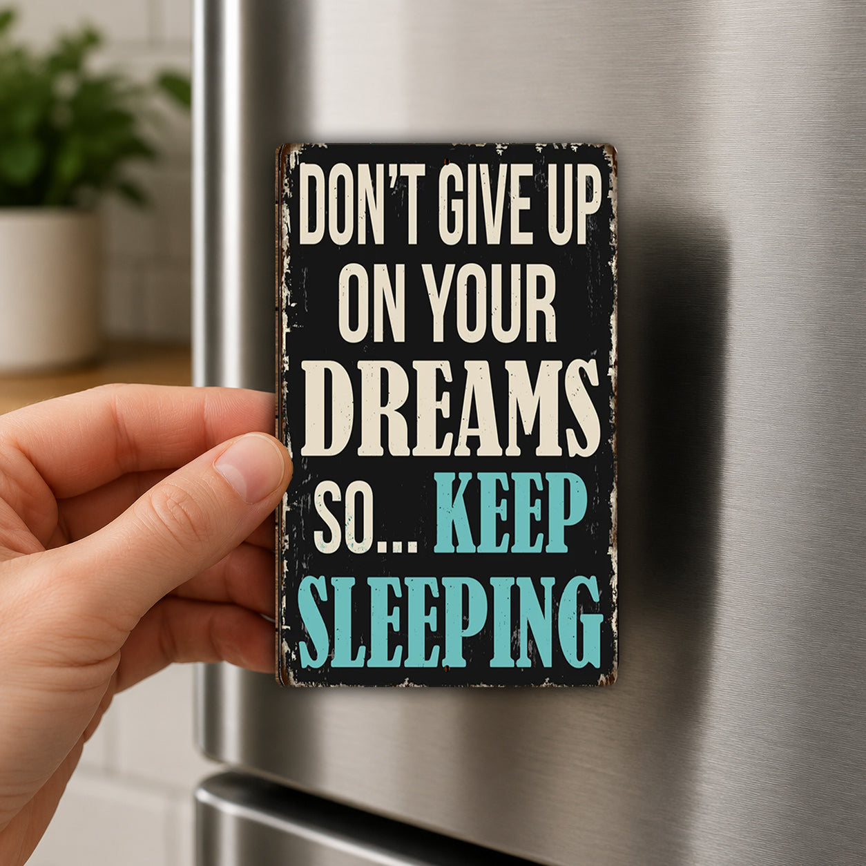 A person's hand holding a sign with the text "DON'T GIVE UP ON YOUR DREAMS SO... KEEP SLEEPING" against a metallic background.