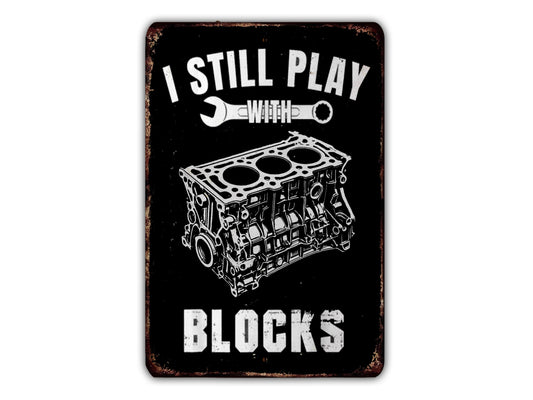 a black and white sign with the text "I STILL PLAY BLOCKS" and an illustration of a wrench and a car engine block.