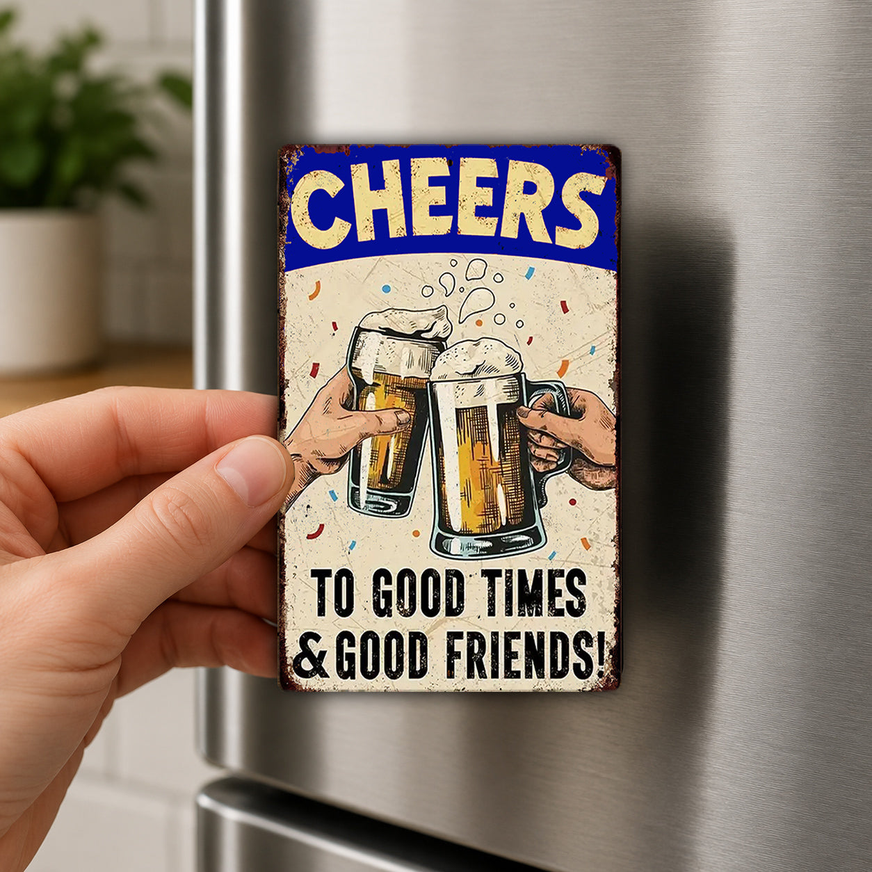 A hand is holding a metal refrigerator magnet with a hand-drawn illustration of two people clinking beer glasses together, accompanied by the text "Cheers to good times & good friends!"