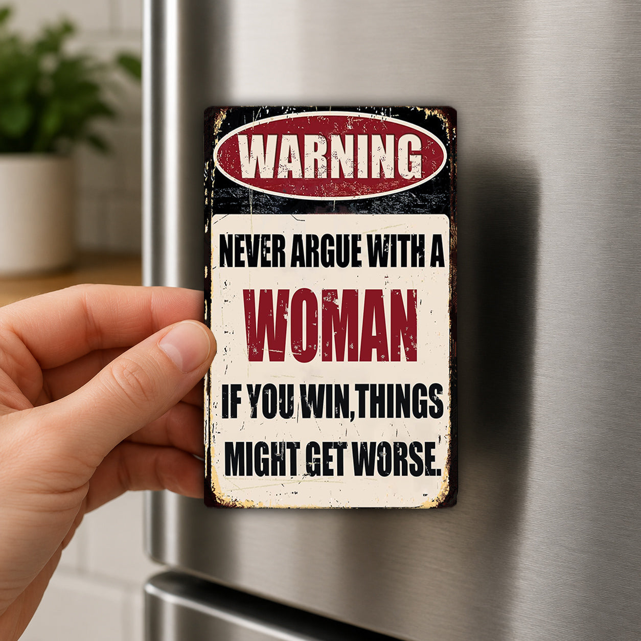 A person's hand is holding a metal sign with the text "WARNING NEVER ARGUE WITH A WOMAN IF YOU WIN, THINGS MIGHT GET WORSE" on it.