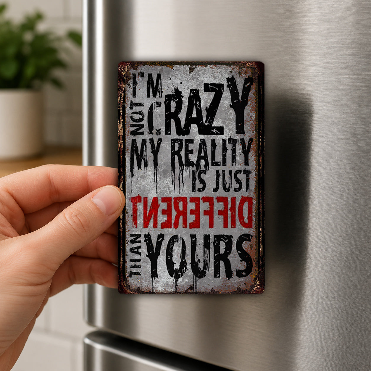 A person's hand is holding a metal sign with the text "I'm not crazy, my reality is just different than yours" on it.