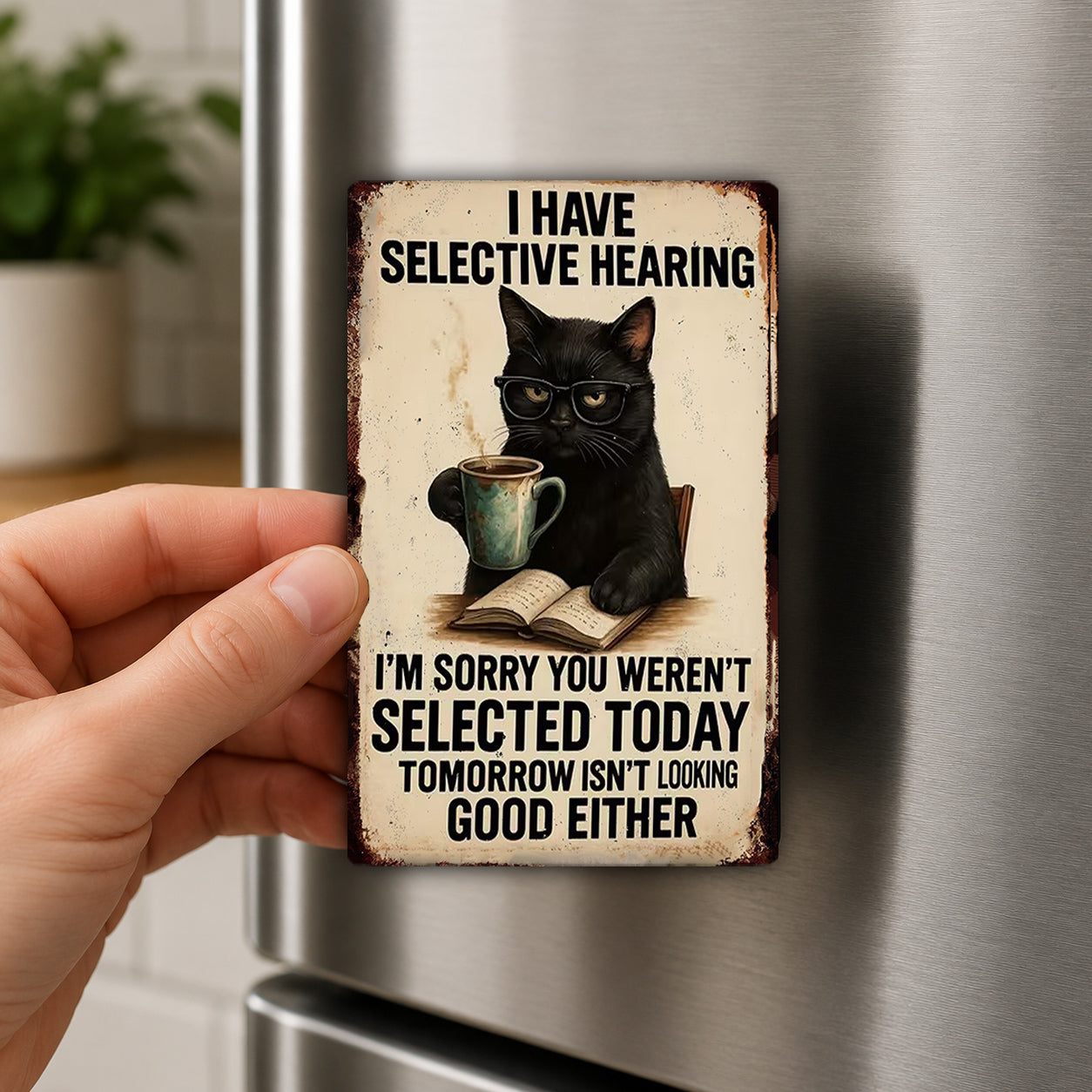 A person's hand holding a metal magnet with a black cat wearing glasses and holding a mug, with text that reads "I HAVE SELECTIVE HEARING I'M SORRY YOU WEREN'T SELECTED TODAY TOMORROW ISN'T LOOKING GOOD EITHER".