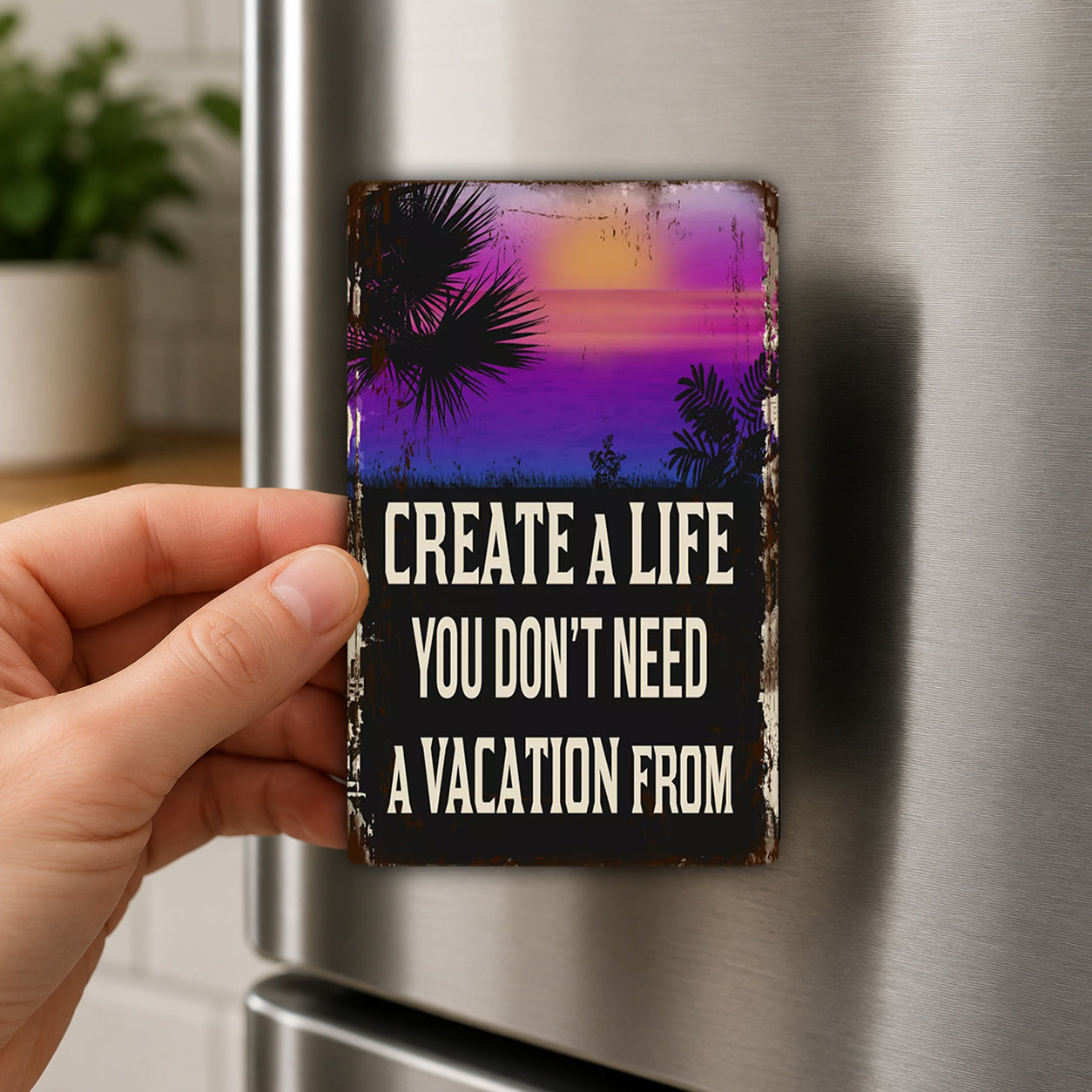 A hand is holding a small, vintage-style magnet with a beach scene and palm trees on it. The magnet has the text "CREATE A LIFE YOU DON'T NEED A VACATION FROM" written on it.
