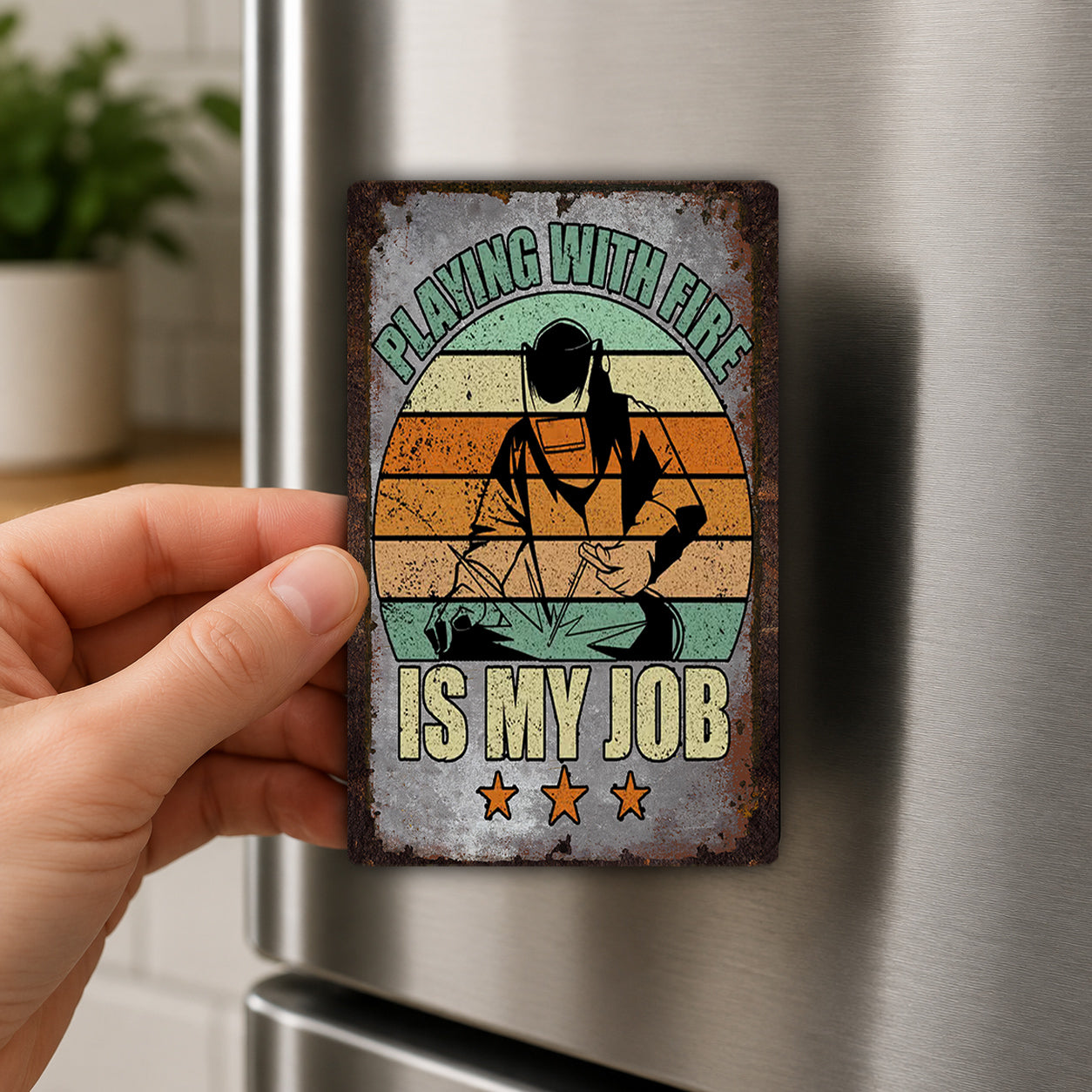 A person's hand holding a metal magnet with a vintage-style design featuring a firefighter and the text "Playing with fire is my job".