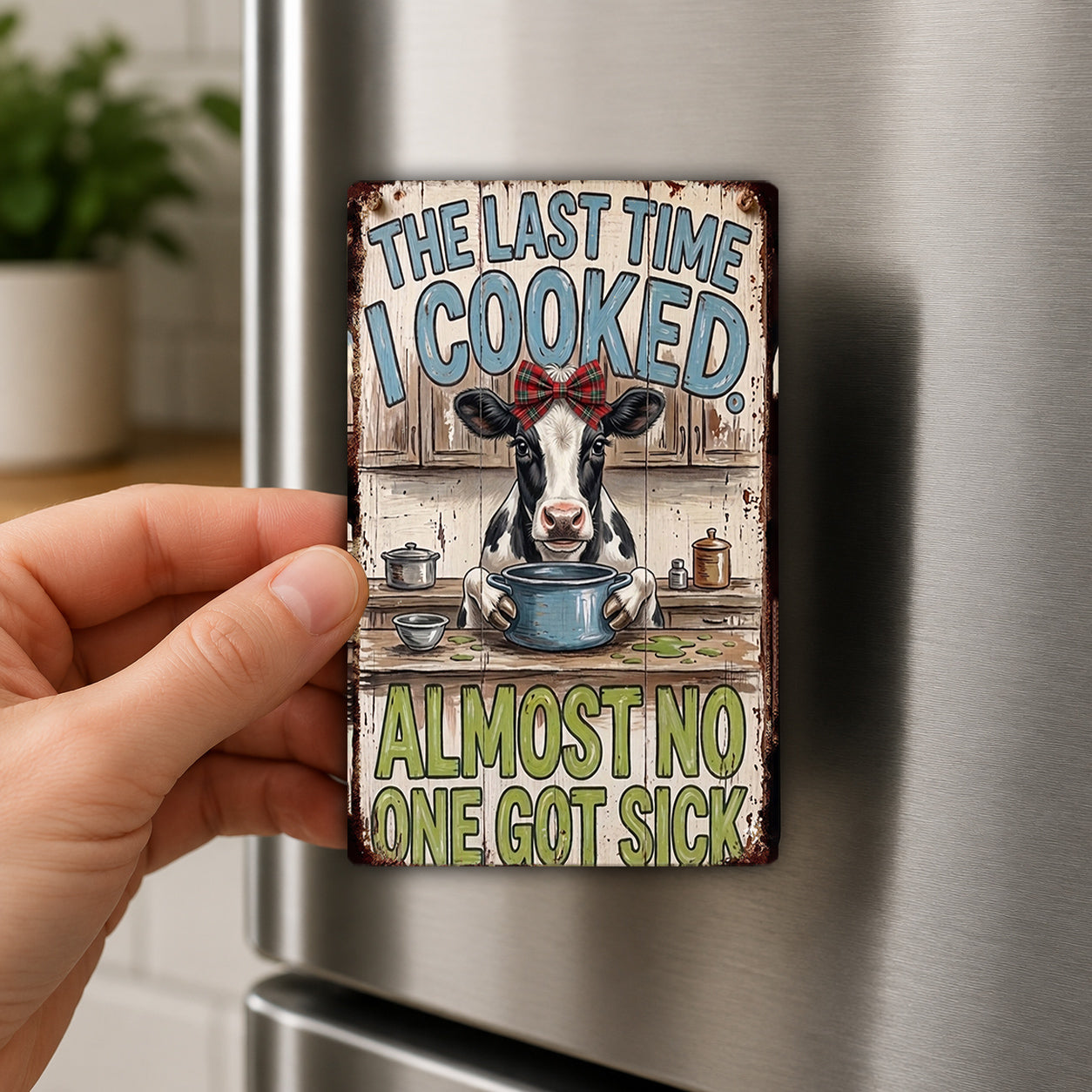 A person's hand holding a metal refrigerator magnet with a cartoon cow and text on it.